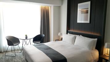 A hotel room with a large bed, two chairs, a small table, a nightstand with a lamp, and a window with curtains.