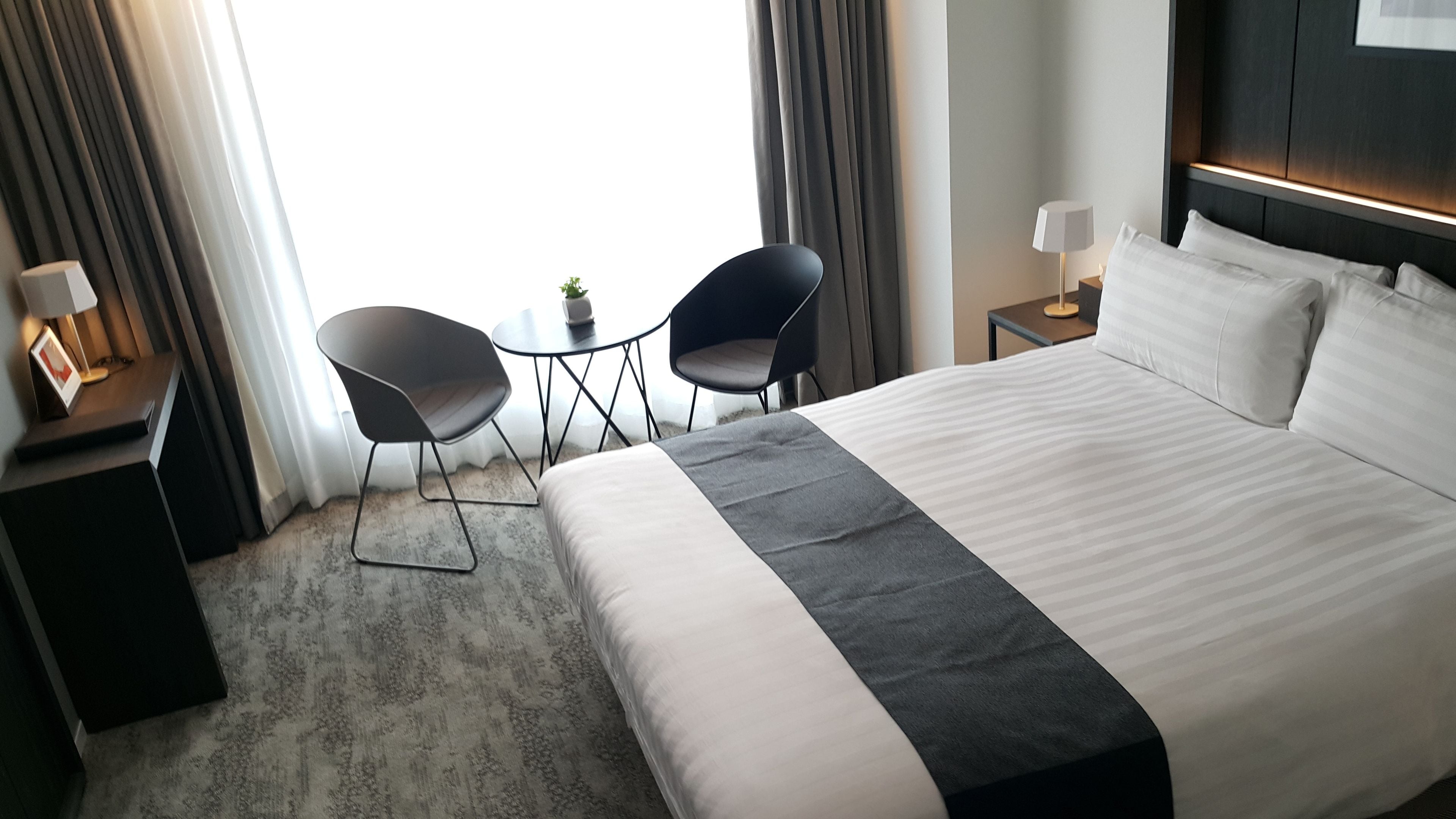 A hotel room with a large bed, two chairs, a small table, a lamp, and a view of the outdoors.