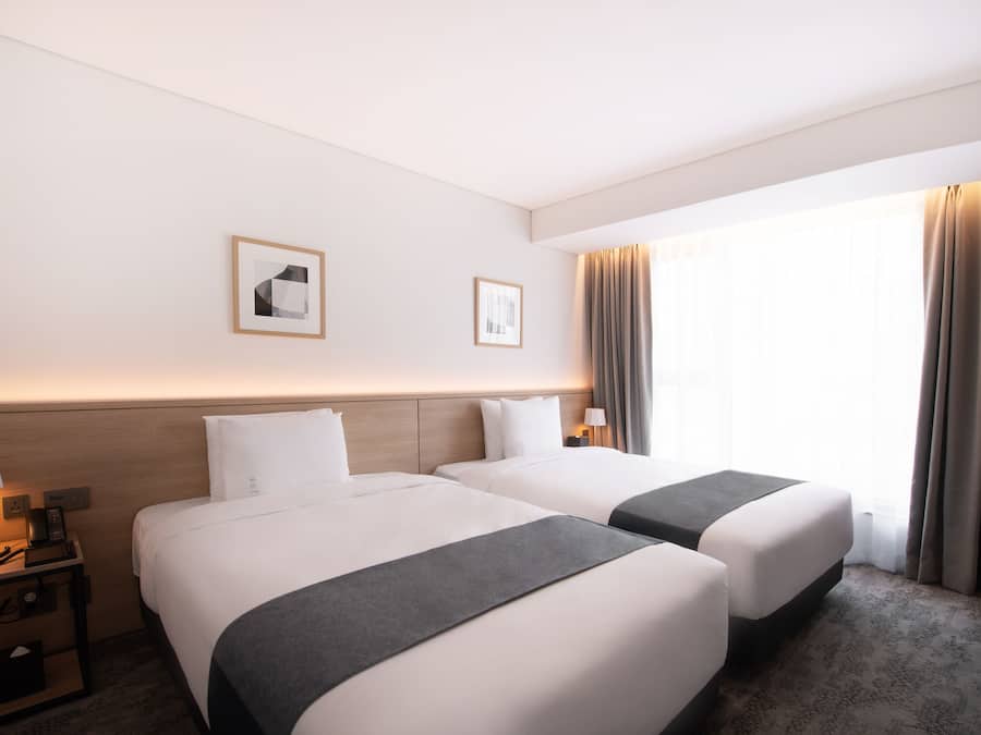 A hotel room with two beds, a nightstand with a lamp, and a window with curtains.