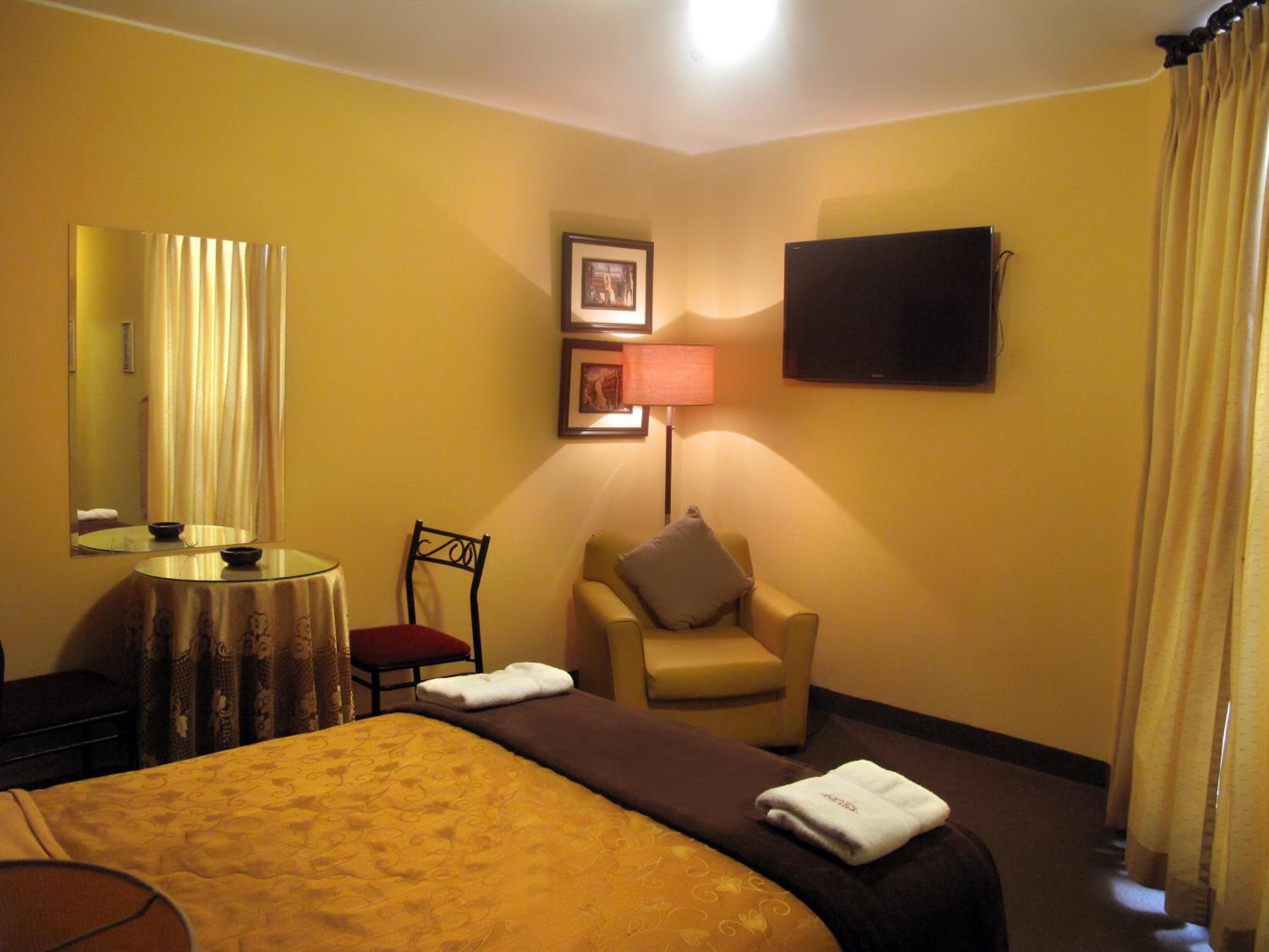 A hotel room with a bed, a chair, a desk with a lamp, a flat-screen TV, and a mirror.