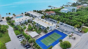 Vista aérea de um hotel à beira-mar, com vários edifícios, quadra de tênis e uma praia ao fundo.