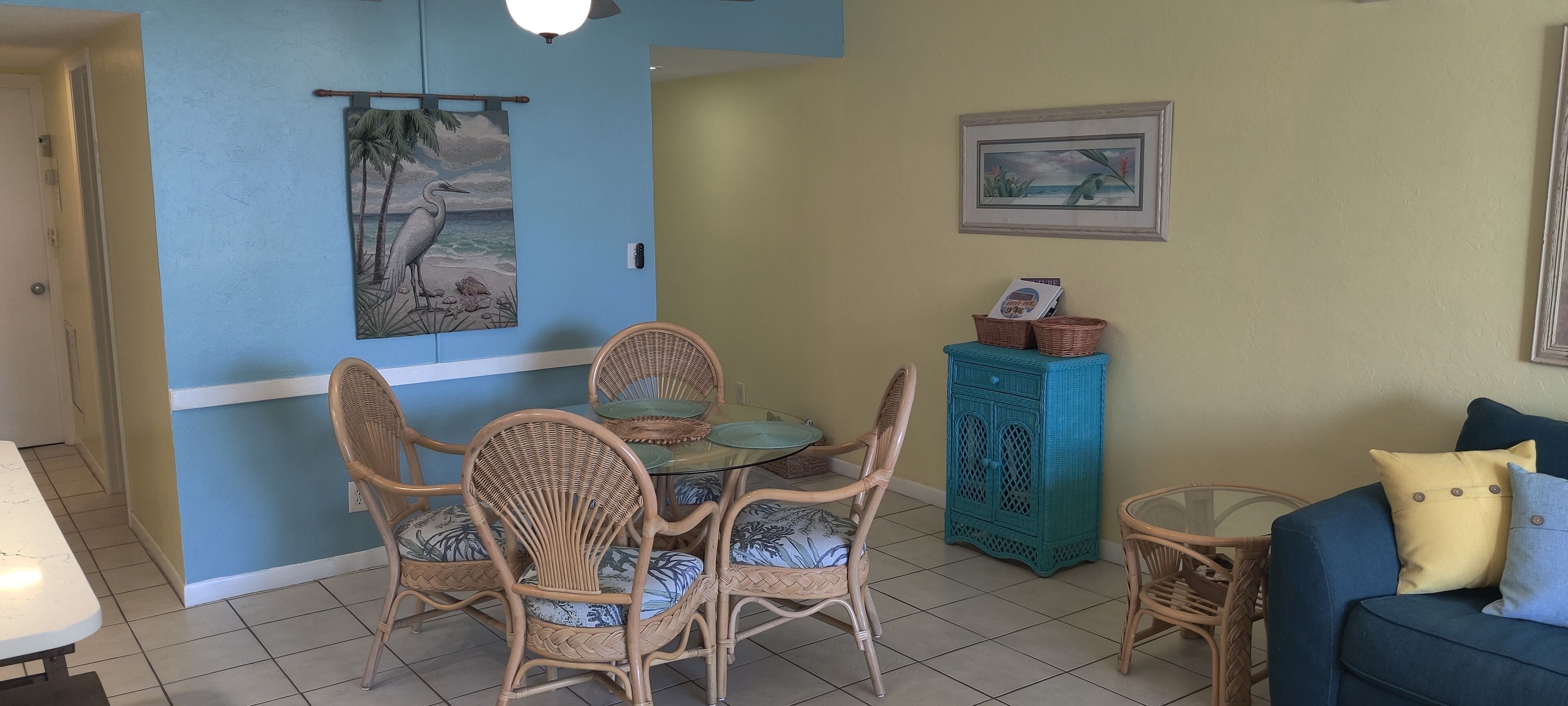 A dining area with wicker chairs and a glass table, a teal cabinet, and a framed picture on the wall.