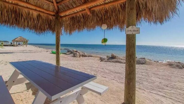 Uma praia com uma estrutura de telhado de palha, uma mesa e uma placa indicando o nome da praia.