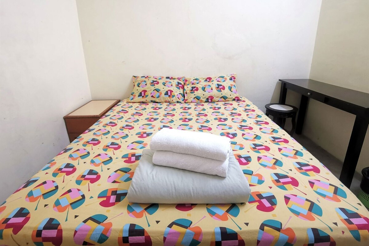 A single bed with a colorful geometric-patterned bedspread, two pillows, a neatly folded white towel, a small wooden nightstand, and a black desk with a chair.