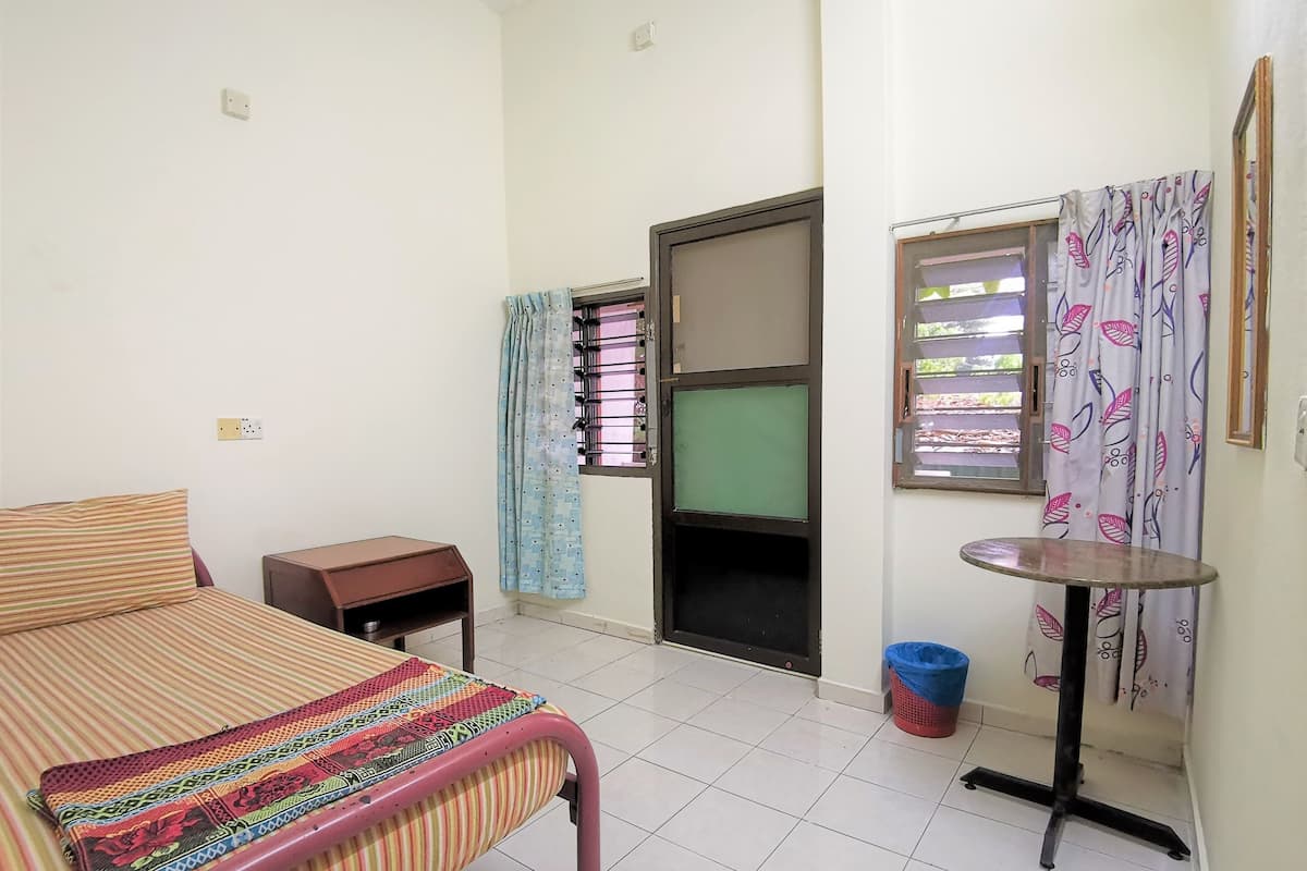 A room with a bed, a small table, a chair, a window with patterned curtains, and a door with a metal grill.