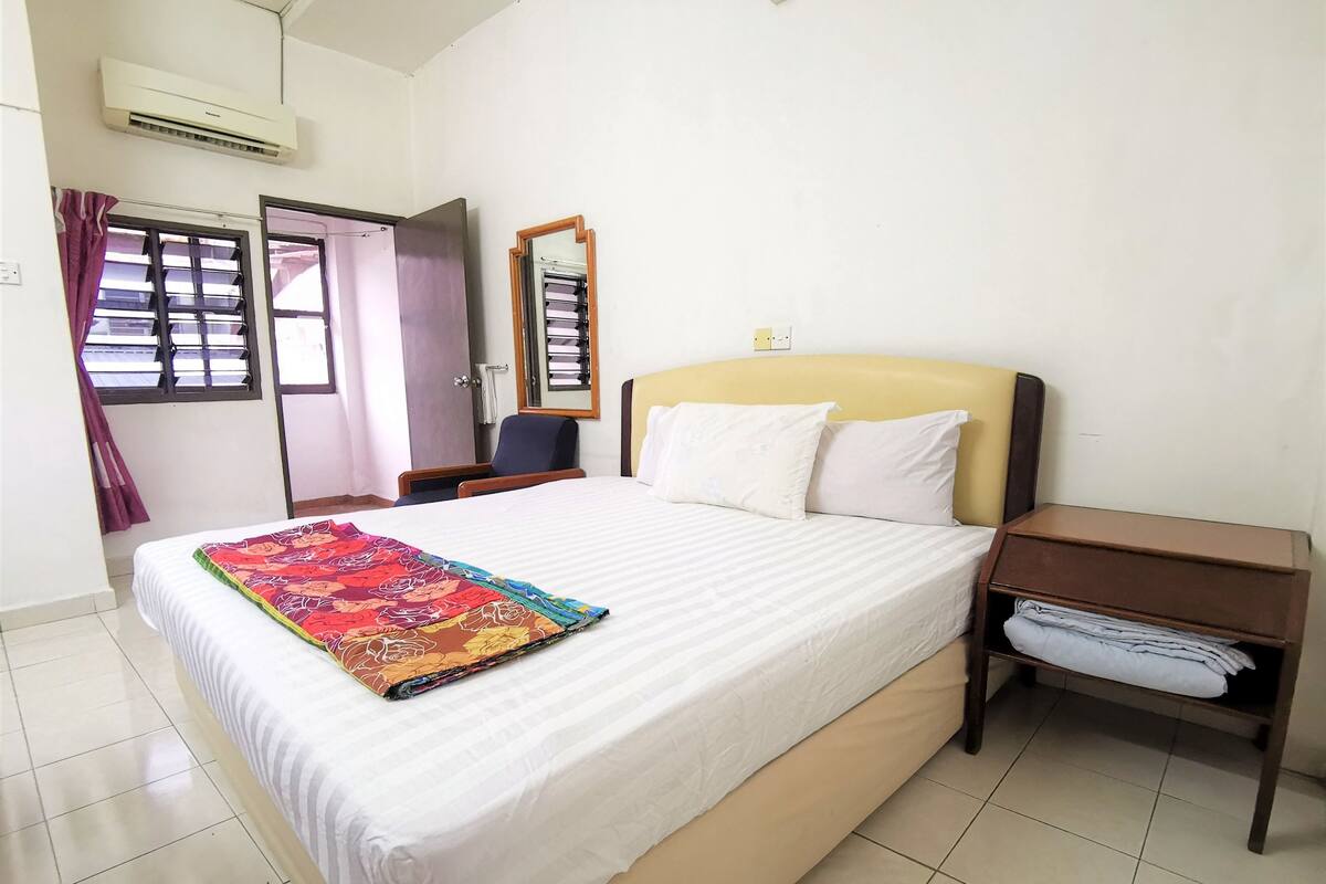 A hotel room with a bed, a nightstand, a chair, a window with curtains, and an air conditioning unit.