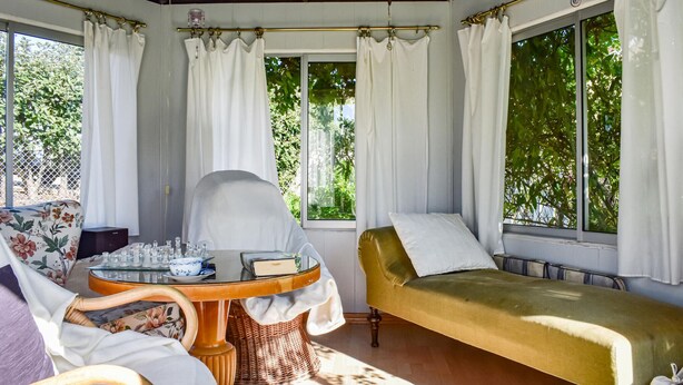 Una habitación iluminada por el sol, con un sofá verde, una mesa redonda con una tetera y una silla con un cojín estampado con motivos florales.