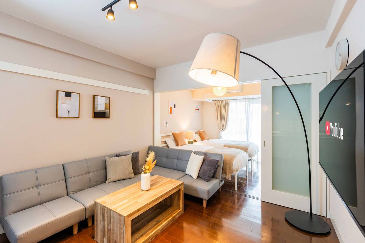 Una habitación de hotel moderna con un sofá gris, una mesa de centro de madera, una cama con ropa blanca, una lámpara de pie grande y una pantalla de televisión mostrando el logotipo de YouTube.