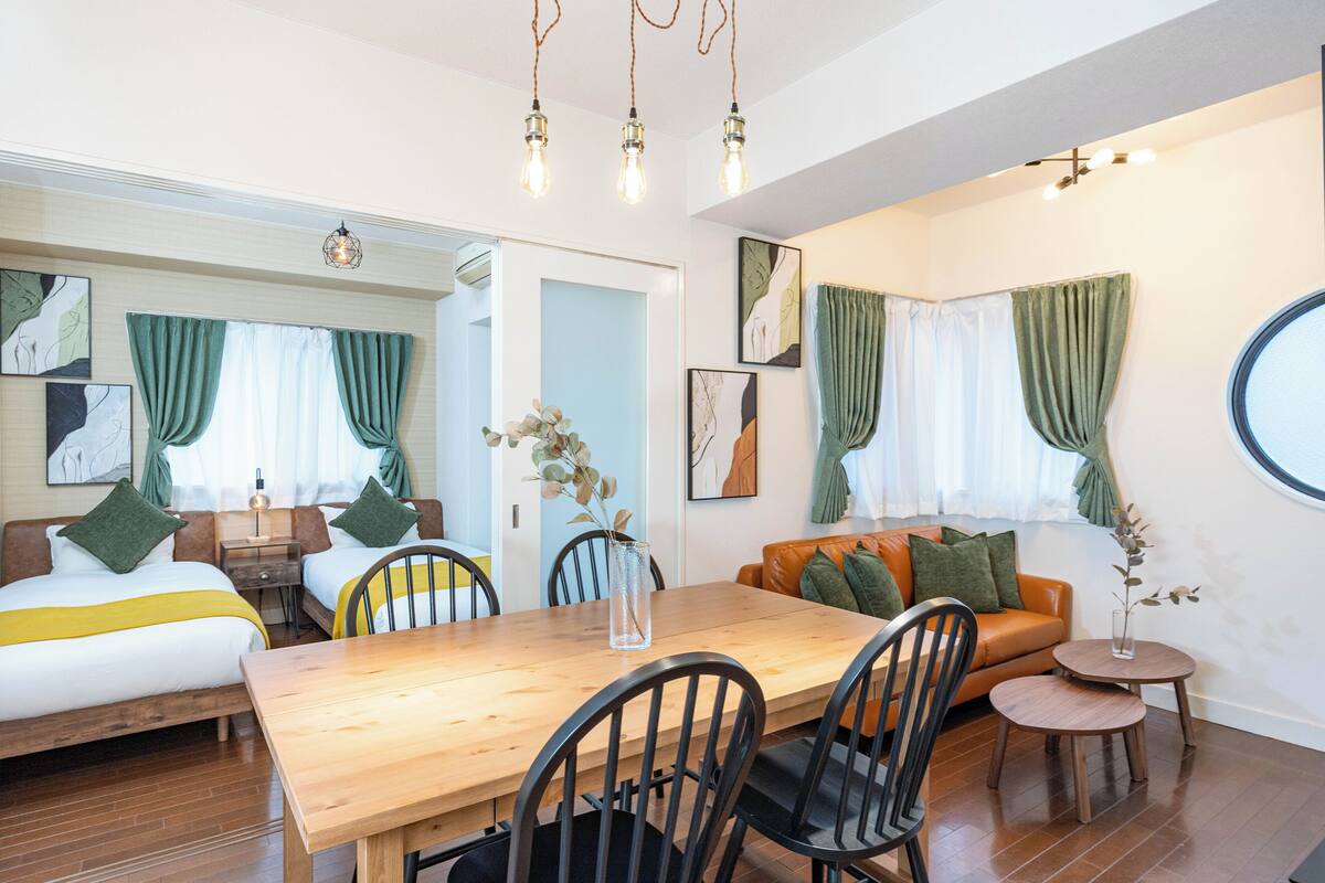 Una habitación de hotel moderna con una mesa de comedor de madera, dos camas con cortinas verdes, un sofá y una pequeña mesa redonda.