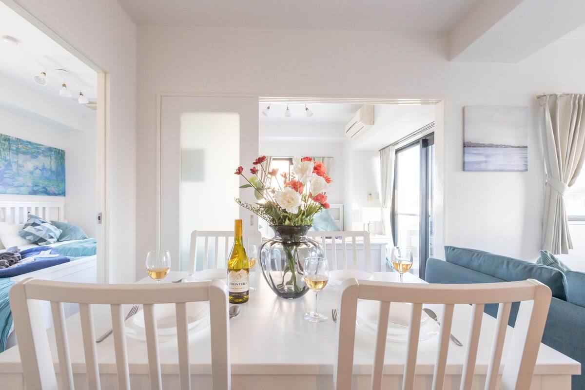 Un comedor con una mesa blanca, dos sillas, un florero con flores y una botella de vino.
