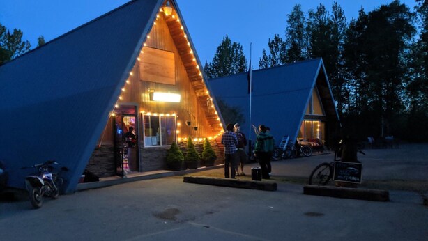 A triangular wooden building with a lit entrance, surrounded by trees. - Talkeetna Inn (Talkeetna)