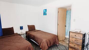 Una camera d'albergo con due letti, un comodino con una lampada, una porta in legno e un comò con cassetti.