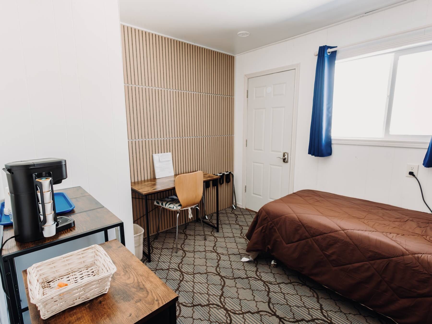 A small hotel room with a bed, a desk with a chair, a coffee machine, a basket, and a window with curtains.