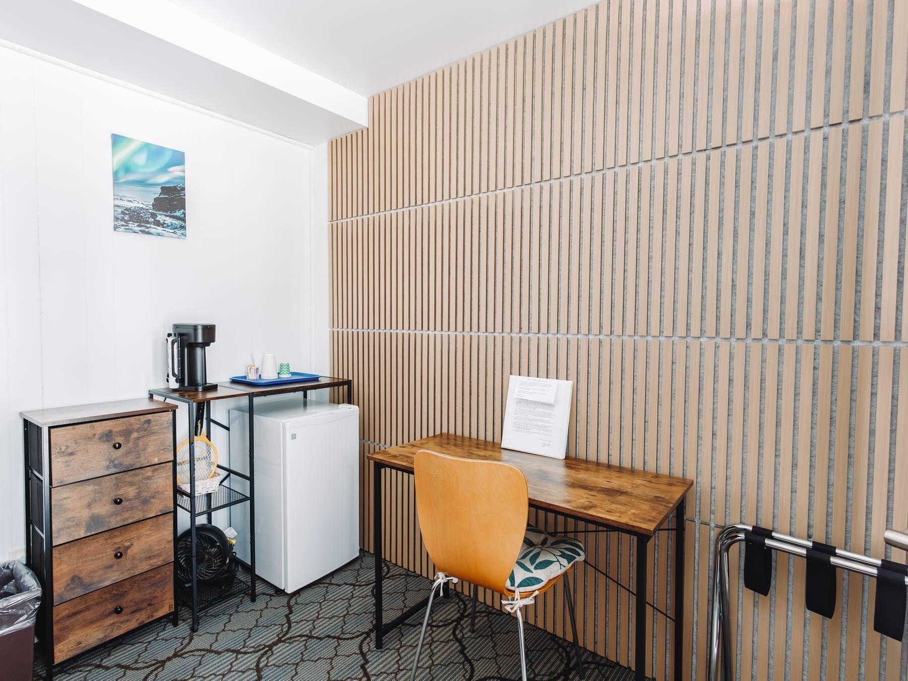 A compact hotel room with a wooden desk, a small refrigerator, a coffee maker, a chair, and a wall-mounted rack.