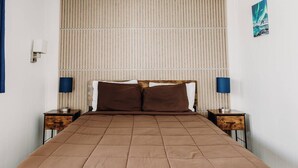A bedroom with a bed, two bedside tables, a wall-mounted light fixture, a vertical striped wall panel, and a framed picture.