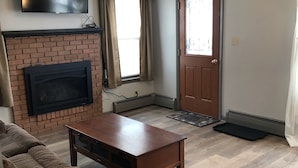 A living room with a brick fireplace, a flat-screen TV mounted above it, a wooden coffee table, a brown sofa, and a wooden door with a glass panel. - Best Location (Cody)