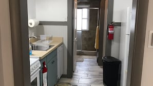 A narrow hallway with a kitchen area on one side, featuring a stove, sink, and cabinets. A fire extinguisher is mounted on the wall, and a trash bin is visible. The floor is tiled with a patterned rug in the hallway. - Best Location (Cody)