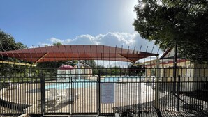 Ett inhägnat poolområde med en överbyggd sittplats och en skylt som anger poolens öppettider.