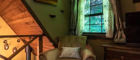 Una habitación con techo de madera, una ventana con cortinas, una silla con cojín y un televisor sobre una mesa de madera.