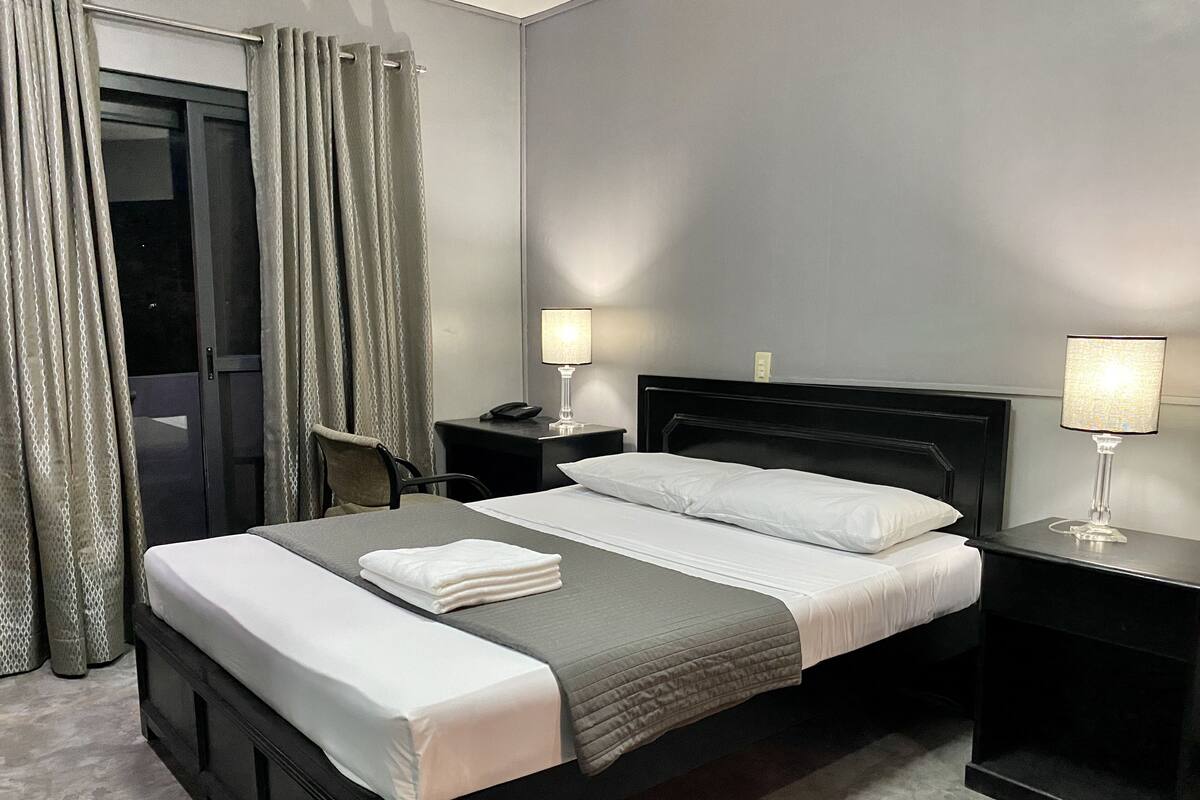 A neatly made bed with white and gray linens, two bedside tables with lamps, a chair, and a view of a balcony with curtains.