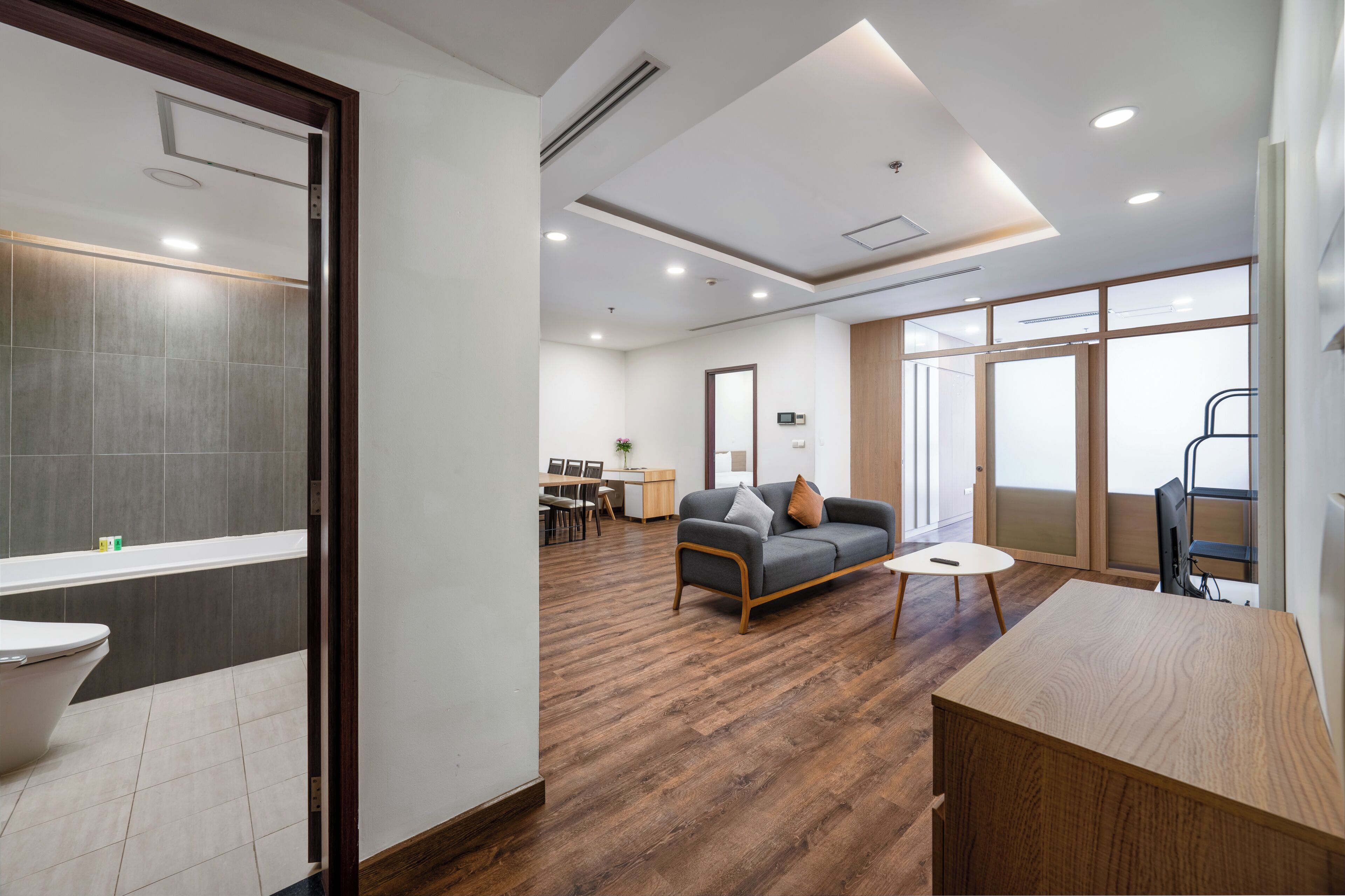 A modern hotel room with a wooden floor, a sofa, a small table, a desk, and a view through large windows.