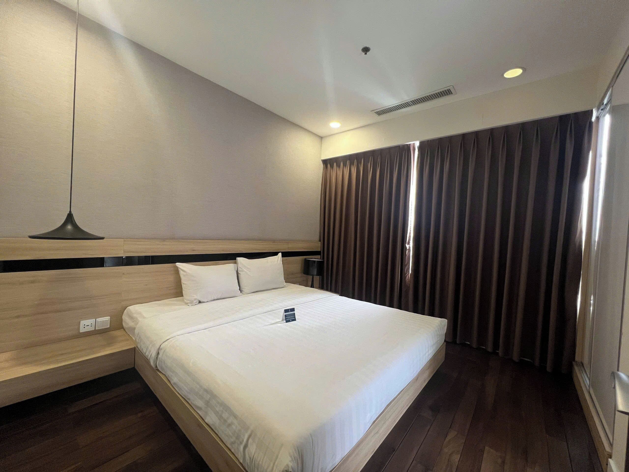 A hotel room with a bed, a bedside lamp, a headboard, a window with curtains, and a wall-mounted light fixture.
