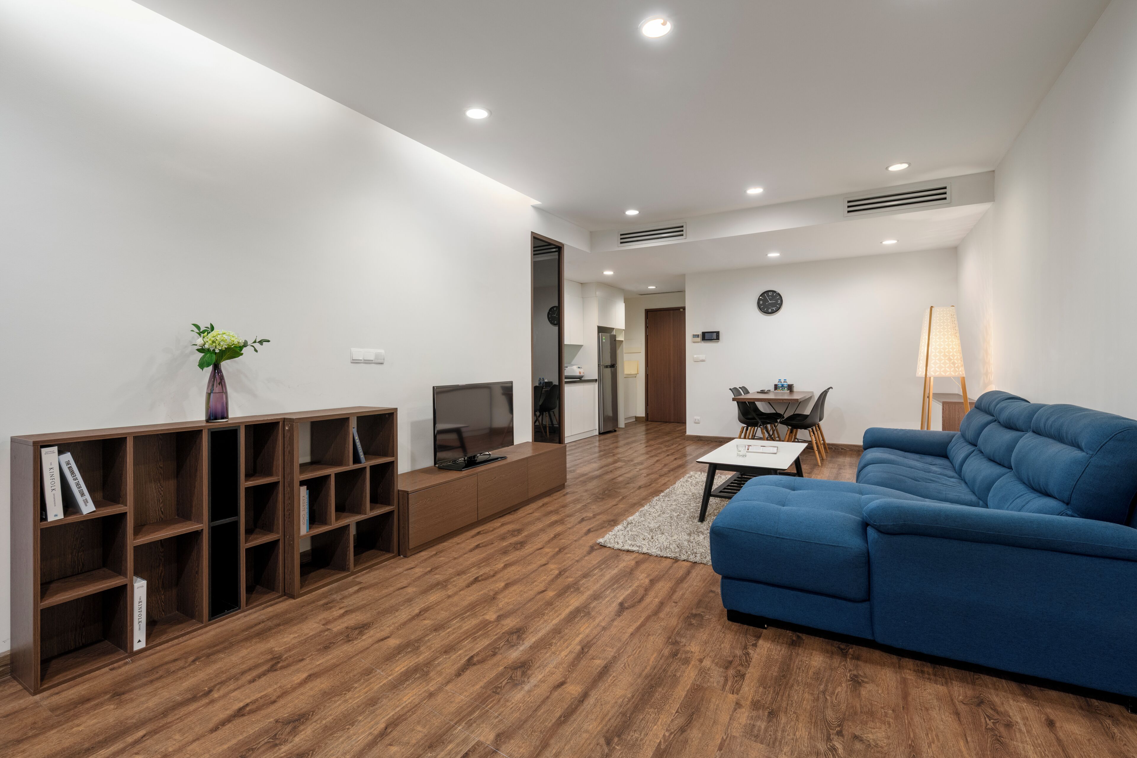 A modern living room with a blue sofa, wooden bookshelves, a TV, and a dining area.