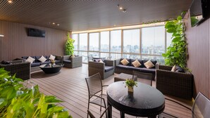 A modern rooftop lounge with wicker furniture, a round table, and large windows offering a city view. - Lancaster Hanoi Service Apartment (Hanoi)