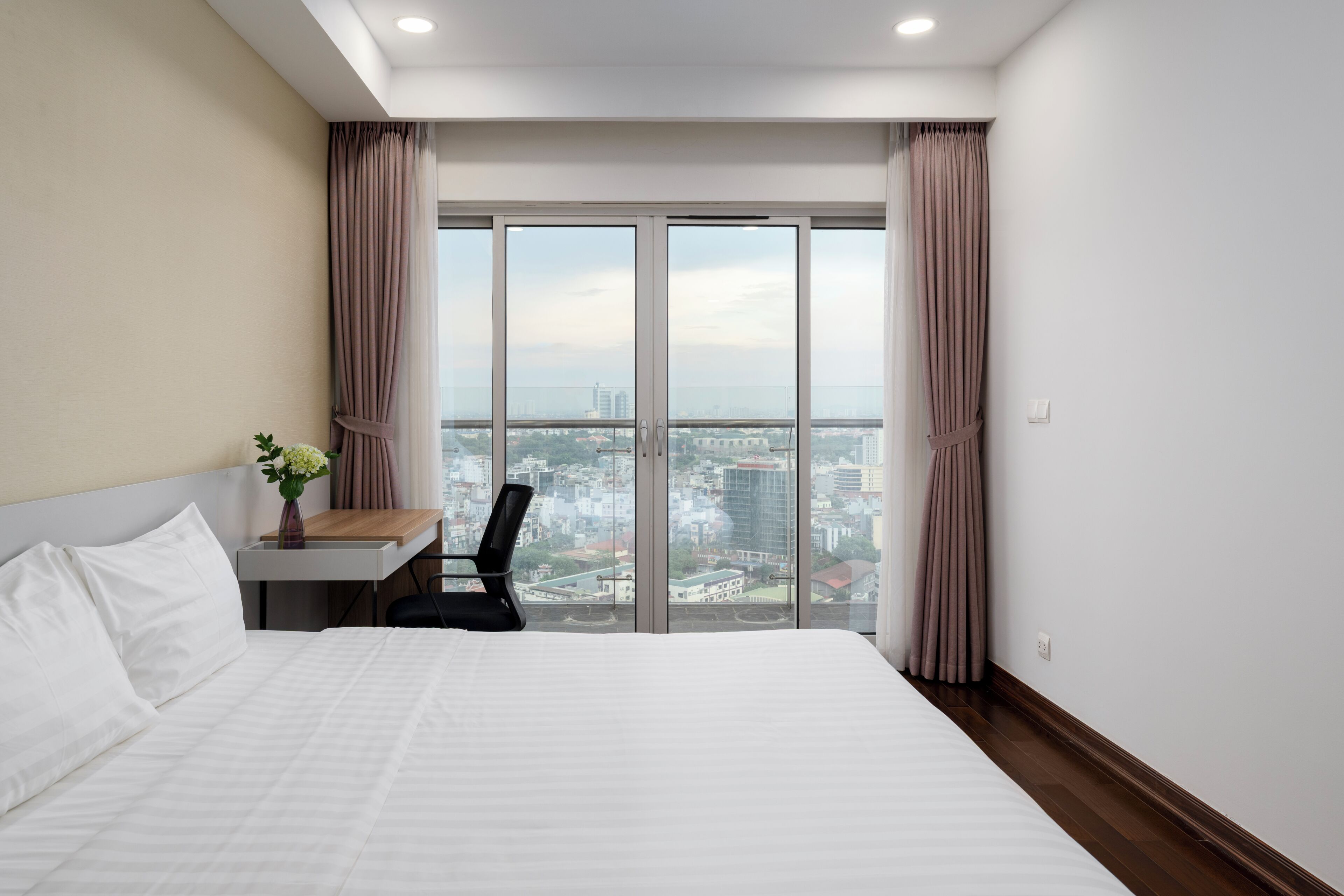 A hotel room with a bed, a desk, a chair, and a view of the city through large windows.