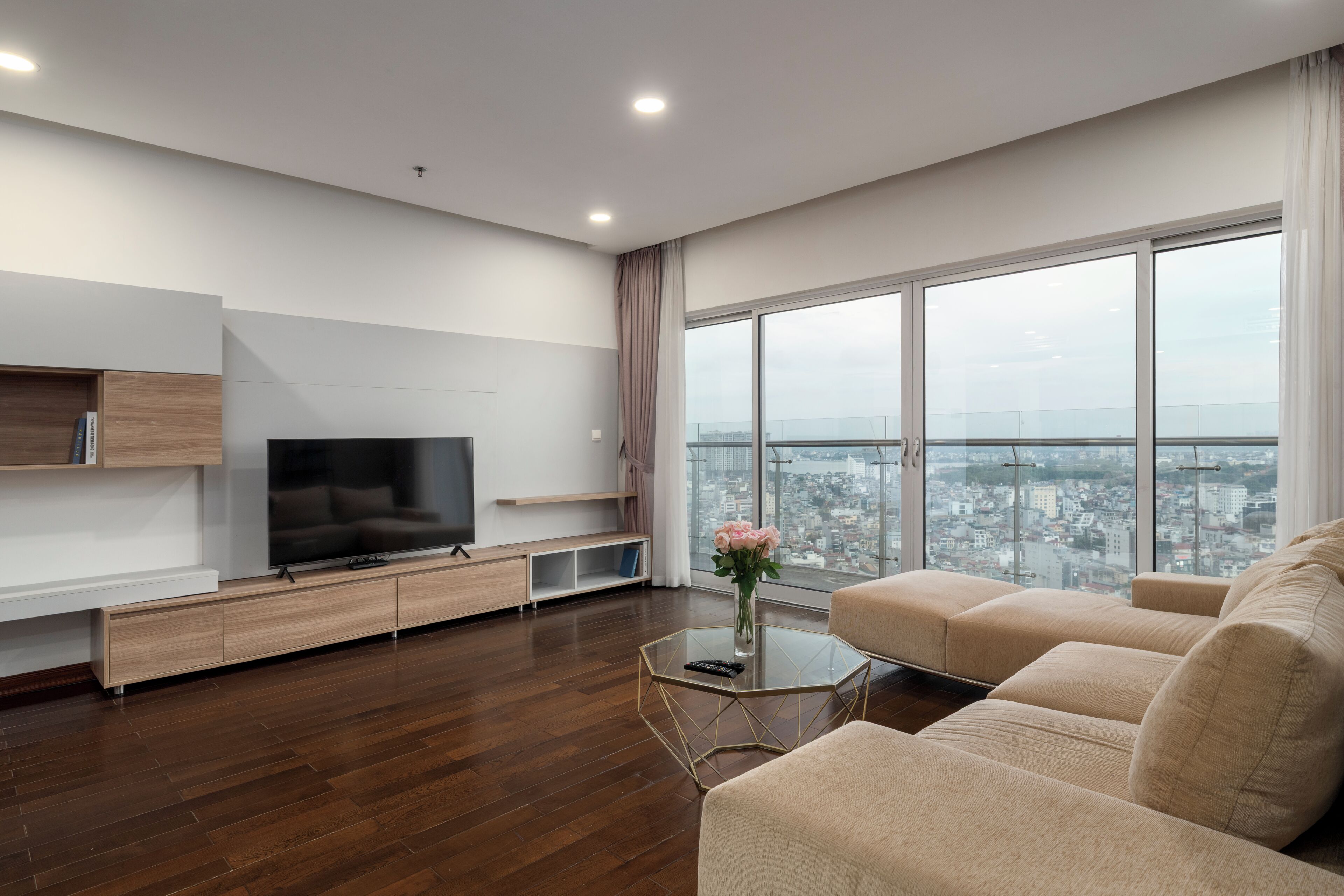 A modern living room with a flat-screen TV, wooden furniture, a glass coffee table, and a large window offering a city view.