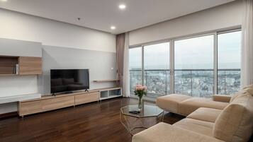 A modern living room with a flat-screen TV, wooden furniture, a glass coffee table, and a large window offering a city view.