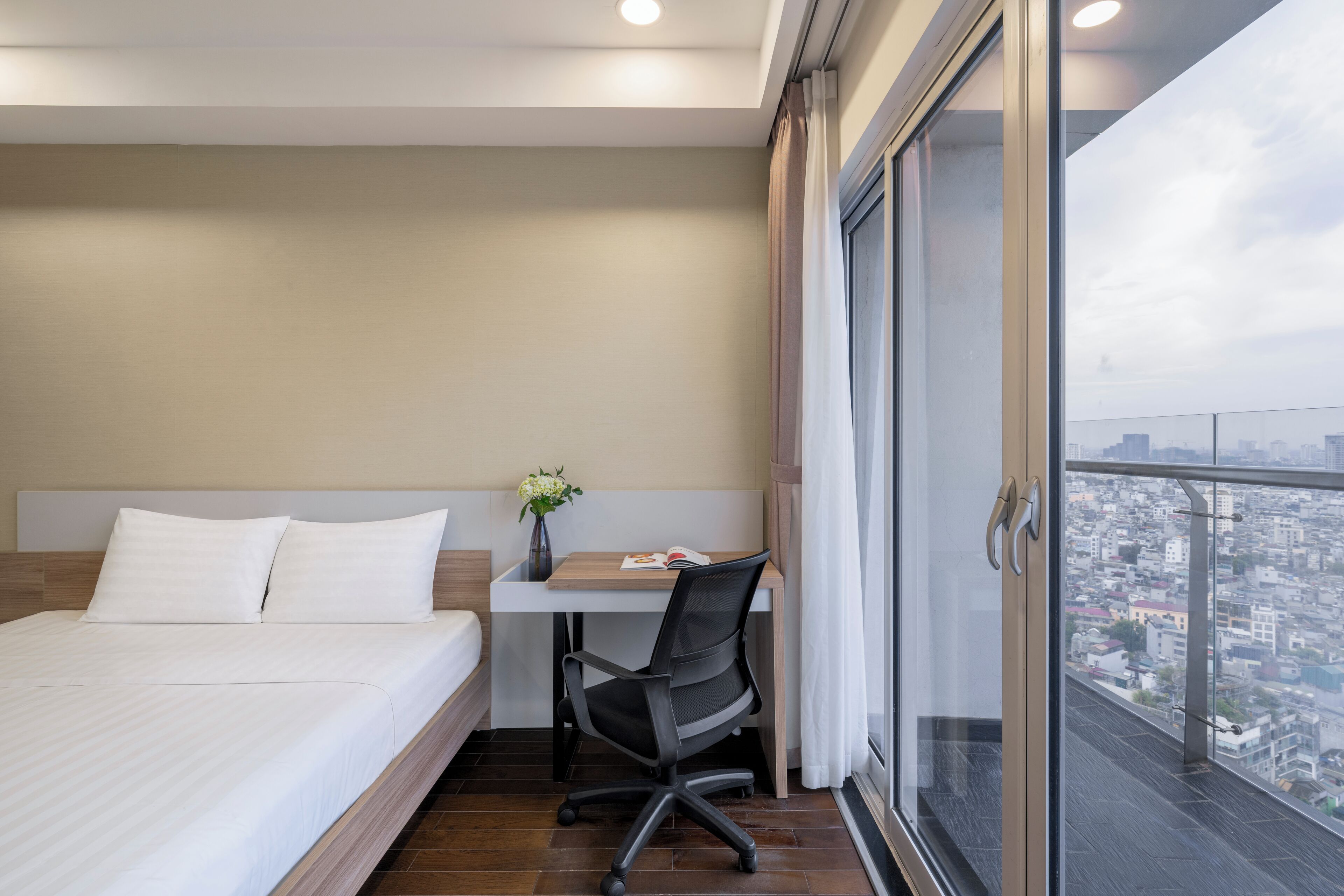 Une chambre d’hôtel équipée d’un lit, d’un bureau, d’une chaise et offrant une vue sur la ville grâce à une grande fenêtre.