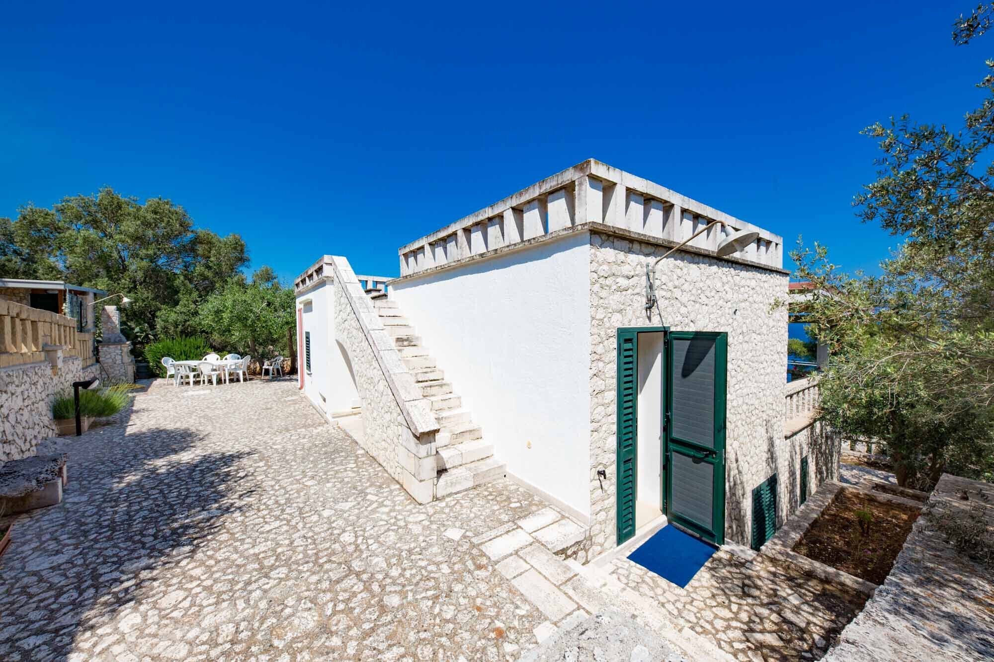 A stone building with a staircase leading to a door, surrounded by cobblestone paths and greenery.