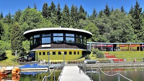 A building with a circular top, a dock leading to a water body, and a forested background.