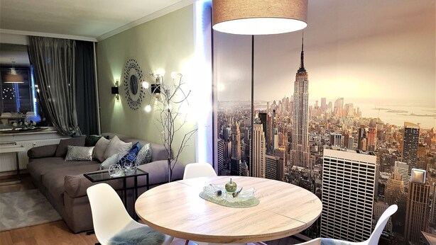 A modern living room with a round wooden table, white chairs, a grey sofa, and a cityscape mural on the wall.
