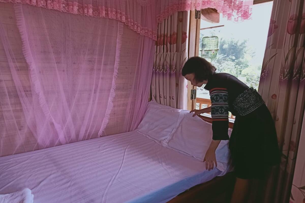 A bed with a pink canopy, a person adjusting the bedspread, and a window with a view of greenery outside.