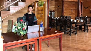 A wooden interior with a staircase, a dining area with tables and chairs, and a person sitting at a desk with a laptop and a floral arrangement. - Sau Meo Homestay (Ta Van)