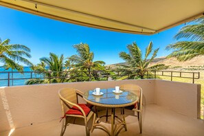 A balcony with a table and chairs, overlooking a beach with palm trees and the ocean. - Summer Deal Oceanfront Maui 2BR Condo with Sunsets and Resort Pool (Wailuku)