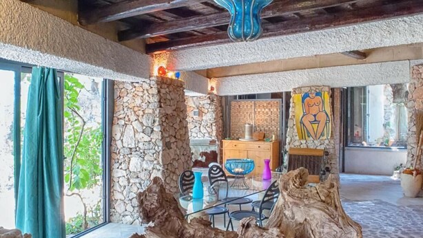 A rustic interior with stone walls, a wooden ceiling, and a dining area with a glass table and chairs.