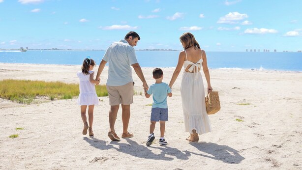 Eine vierköpfige Familie hält Händchen am sandigen Strand, im Hintergrund ein klarer Himmel und ruhiges Meer.