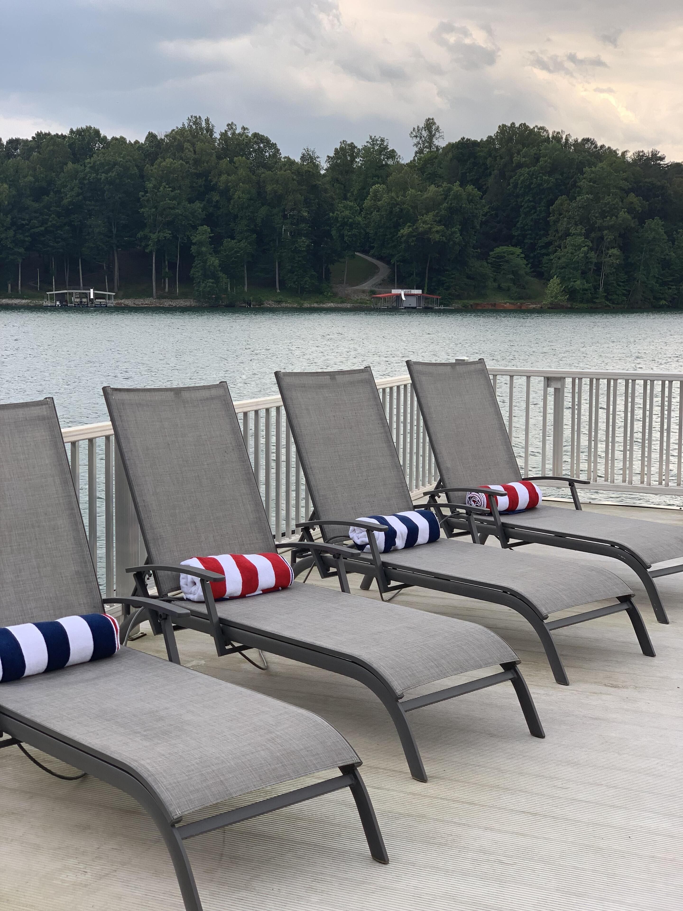 A row of lounge chairs with gray fabric and black metal frames, arranged on a deck with a view of a lake and forested area in the background.