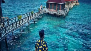 A man in a yellow and black patterned shirt and orange shorts standing on a glass bridge overlooking a clear blue sea with overwater bungalows. - Lato Lato Resort (Semporna)