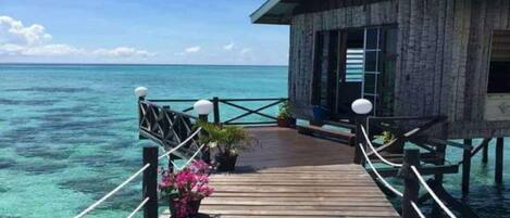 A wooden overwater bungalow with a deck leading to the water, surrounded by clear blue ocean and sky.