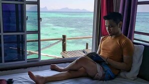 A person sitting on a bed with a view of the ocean through a window. - Lato Lato Resort (Semporna)