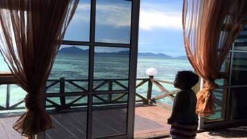 A child standing by a large window with curtains, overlooking a wooden deck and the sea.