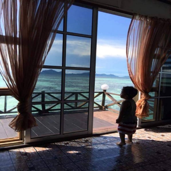 A child standing by a large window with curtains, overlooking a wooden deck and the sea. - Lato Lato Resort (Semporna)