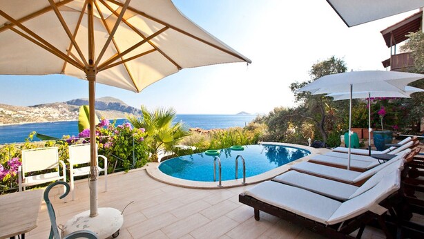 Ein Poolbereich mit Liegestühlen und Sonnenschirmen mit Blick auf eine malerische Landschaft aus Meer und Hügeln.