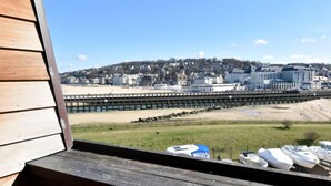 Vista da un pontile di legno su una spiaggia, un molo e una cittadina con edifici e colline sullo sfondo.