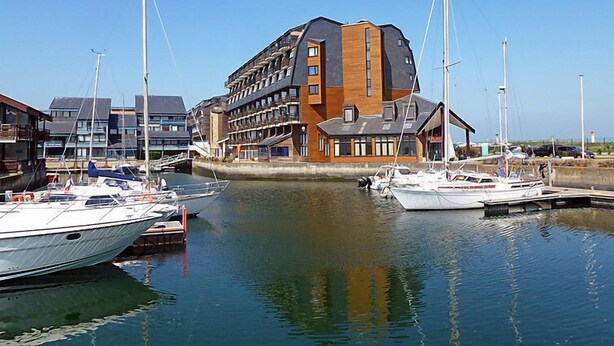 Una marina con diverse imbarcazioni ormeggiate, un edificio moderno con numerosi piani e balconi, e un cielo azzurro limpido.