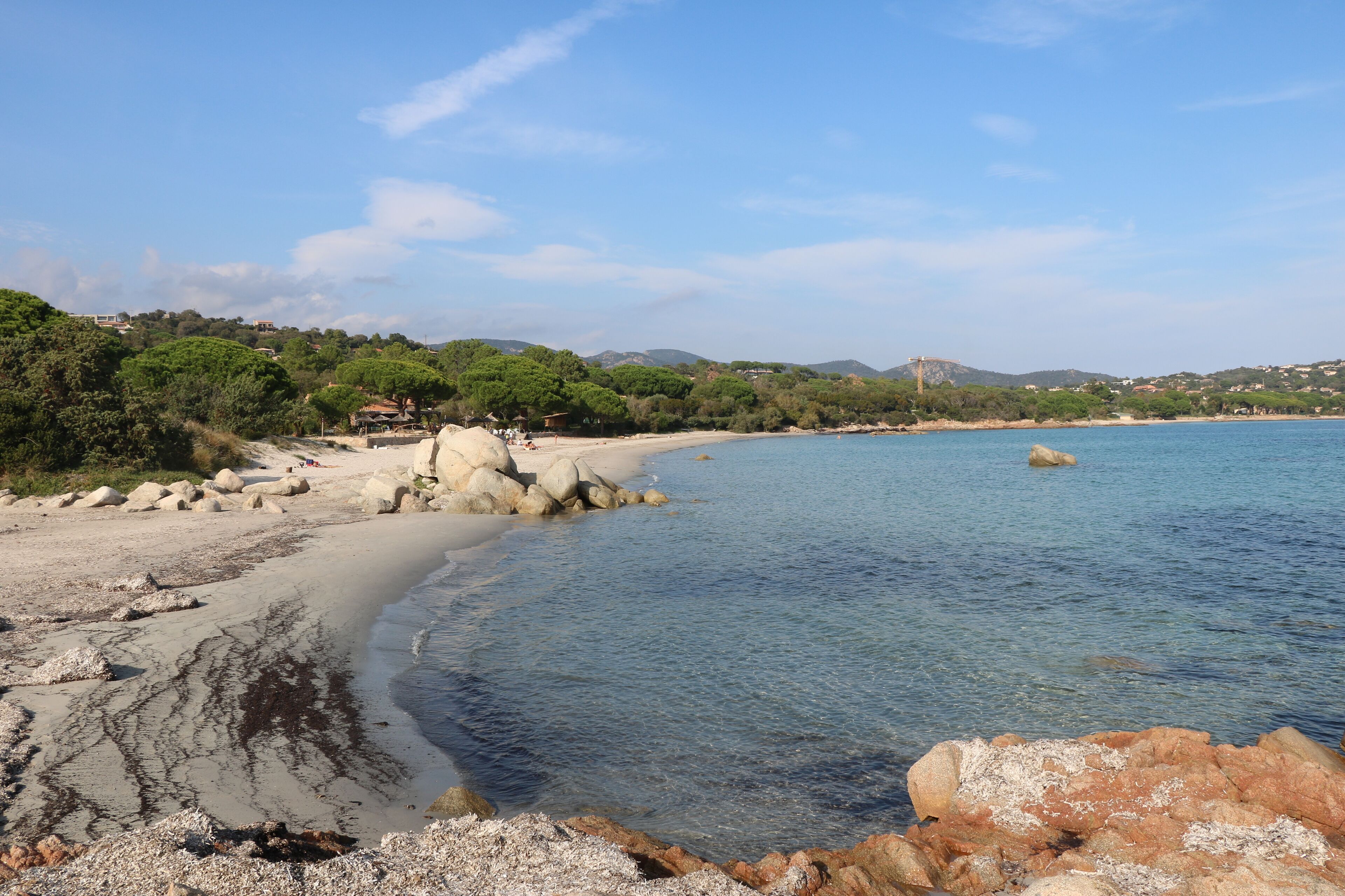A coastal beach with clear water, rocky shore, and lush greenery.